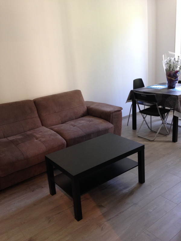 Une pièce de vie lumineuse avec un grand canapé au centre. Devant le canapé, il y a une table basse noire rectangulaire. Sur la droite, on distingue une table à manger avec des chaises.